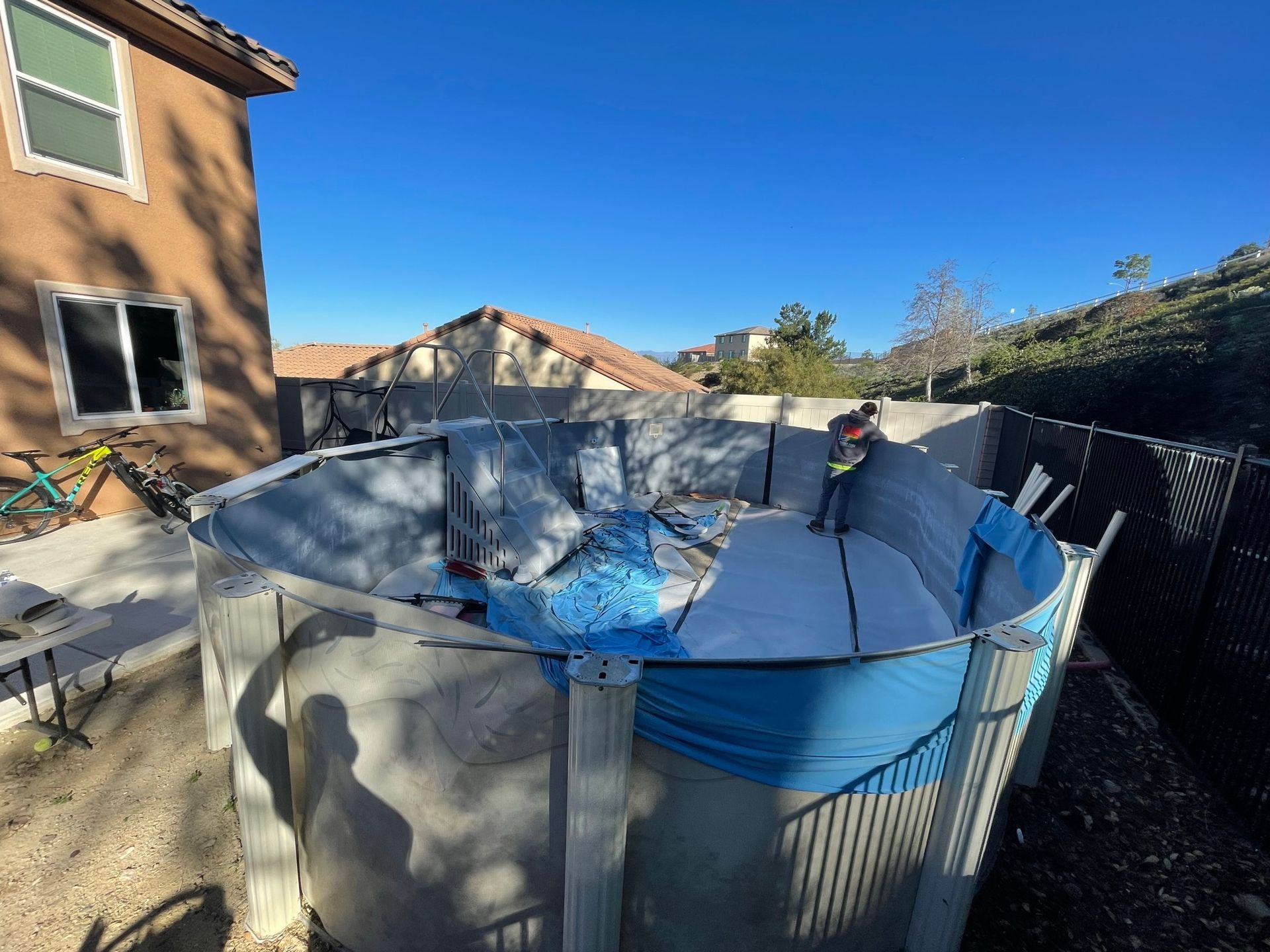 Above-ground pool in a backyard with blue liner, surrounded by a house, fence, and clear sky.