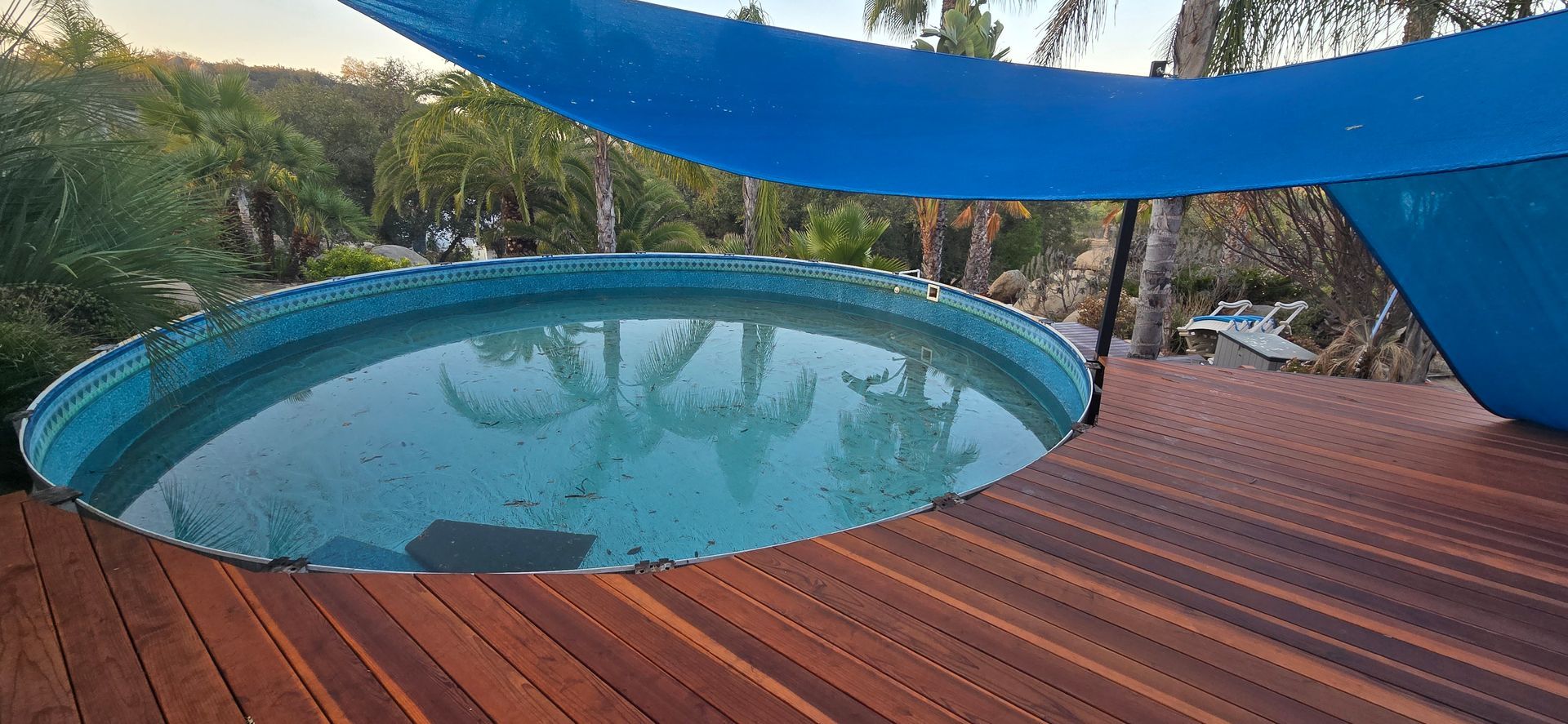 Round pool on a wooden deck under blue shade sails in a wooded backyard