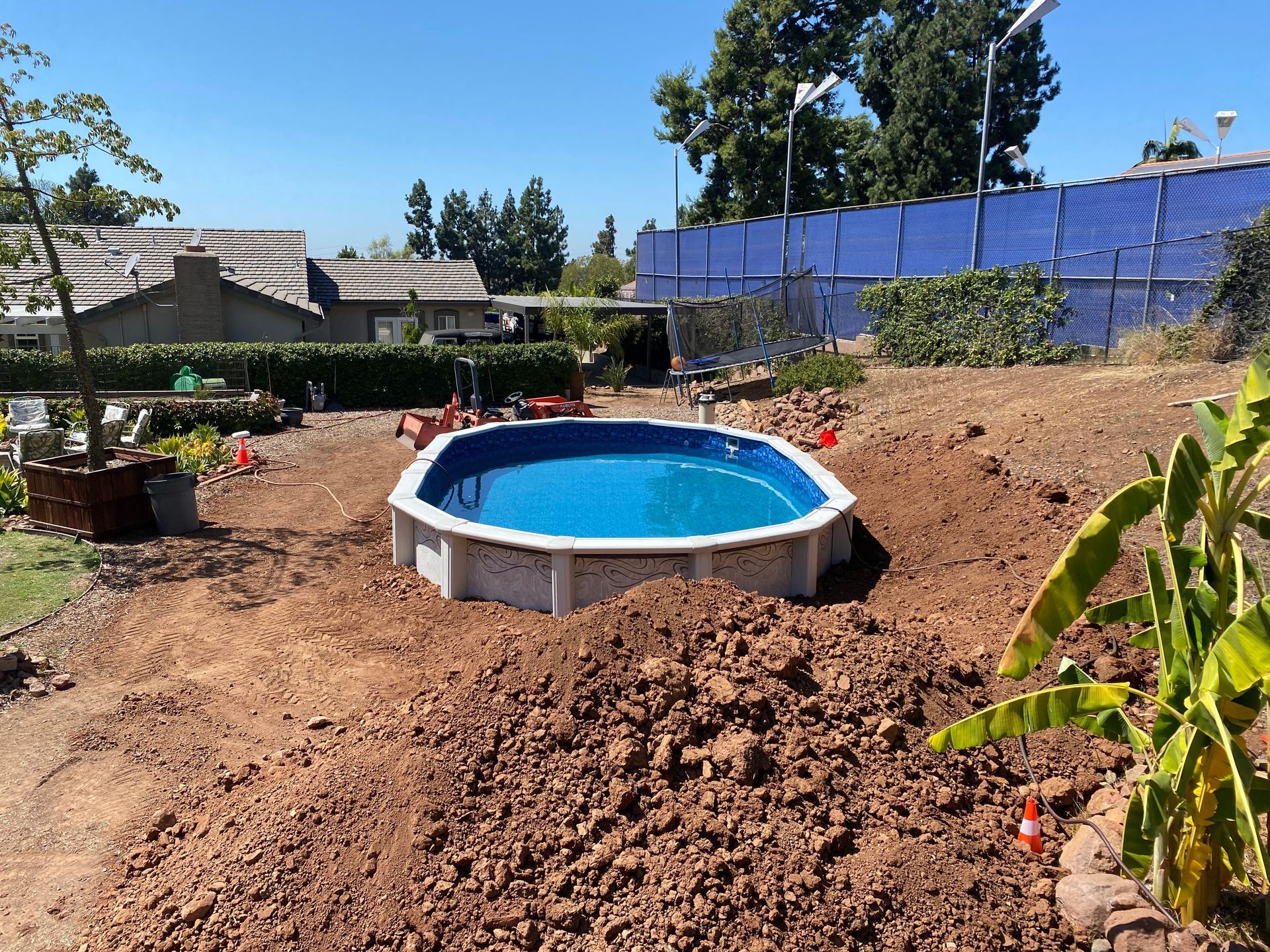 Backyard with above-ground pool, dirt landscaping, and blue fence under clear sky