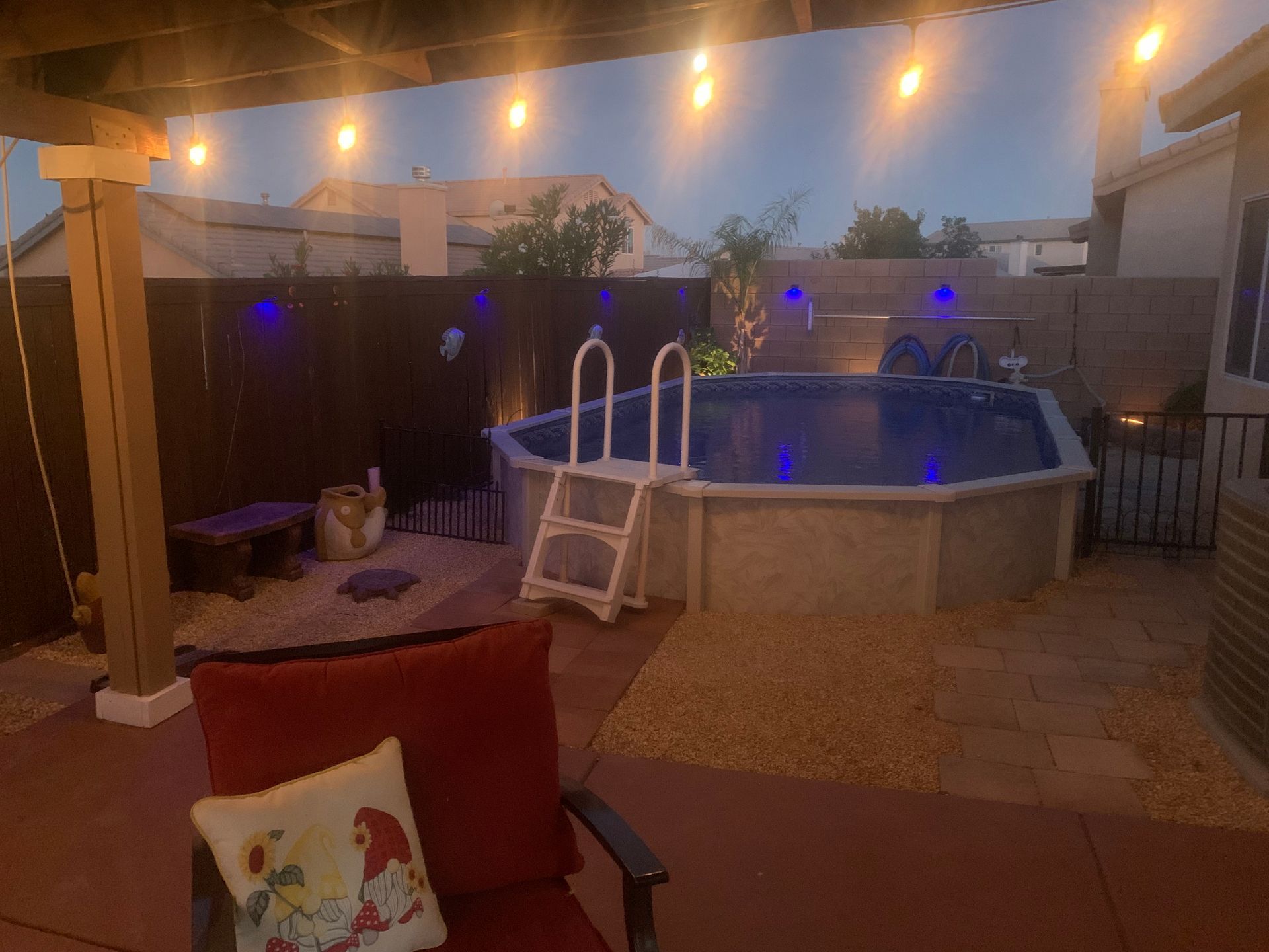 Backyard patio at dusk with lit string lights, above-ground pool, and red chair in foreground