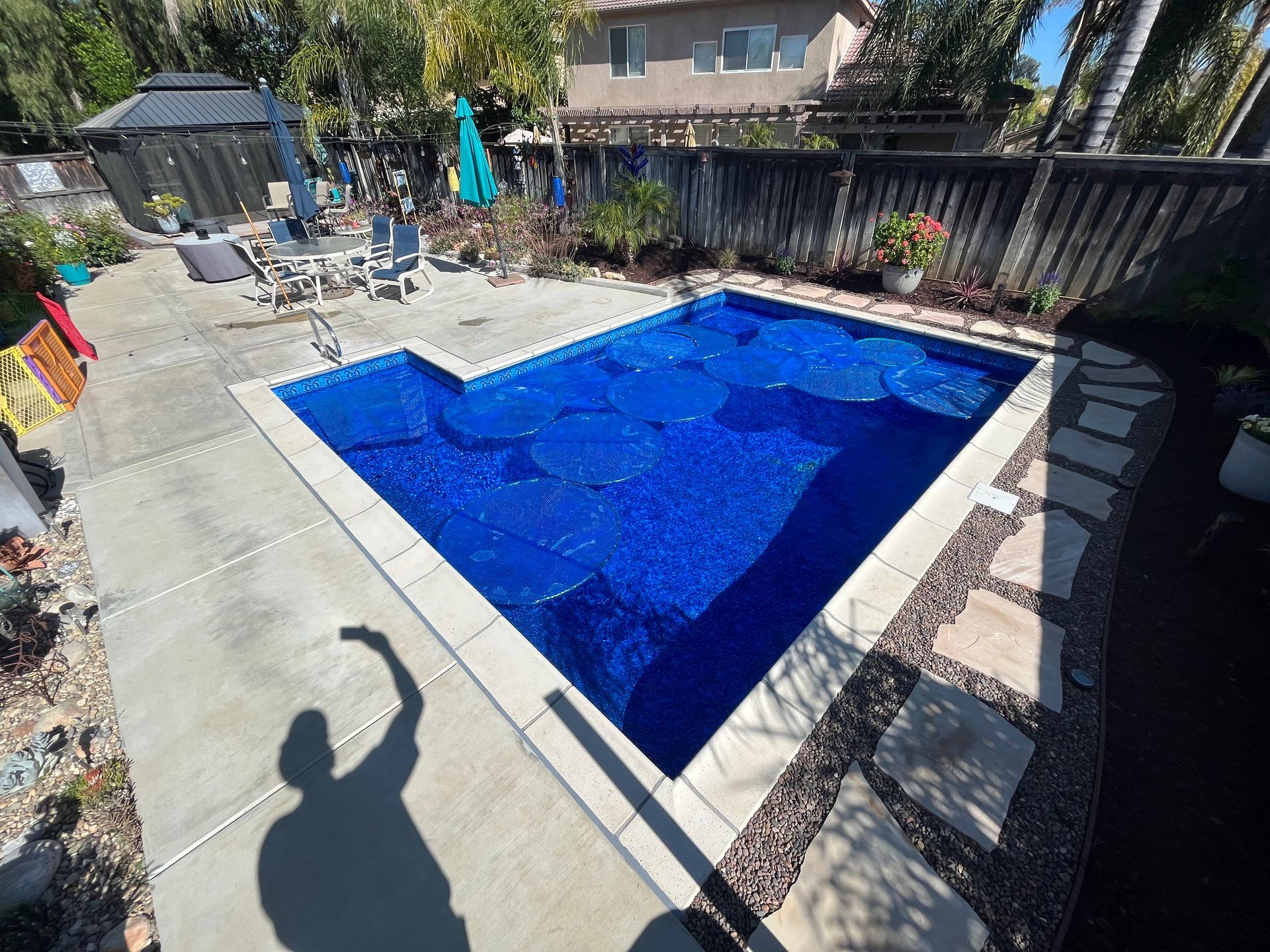 Backyard swimming pool with bright blue water, patio furniture, and a person’s shadow on the deck