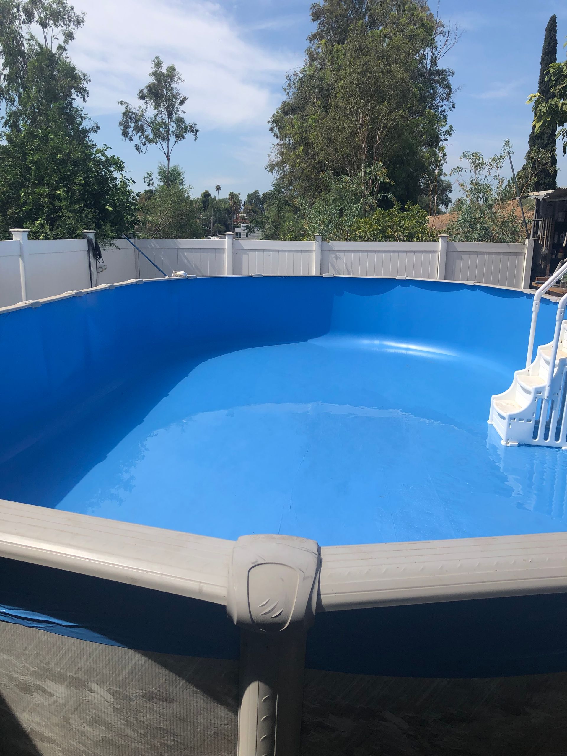 Empty blue backyard swimming pool with white fence and trees in the background