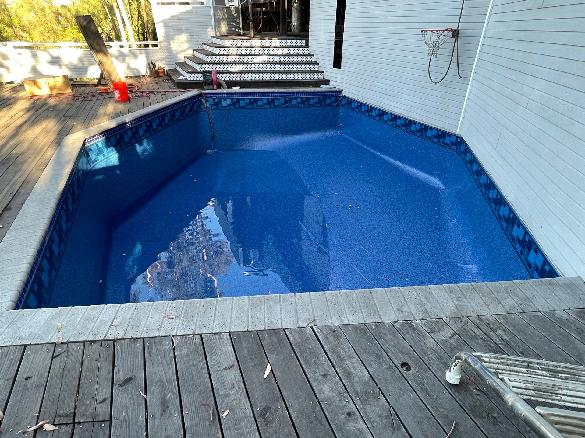 Empty blue backyard swimming pool with a wooden deck and steps leading down from a house.