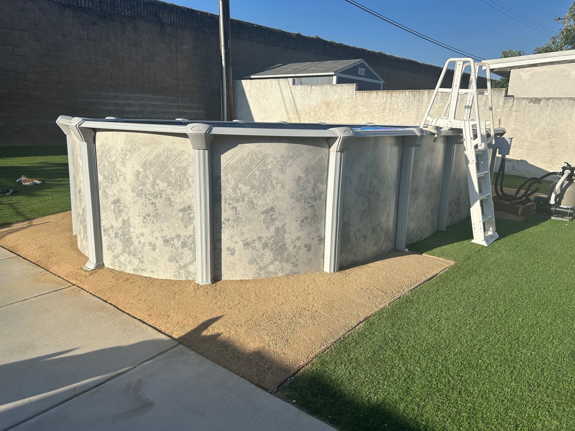 Above-ground pool beside a lawn and patio, with a ladder and white fence in a backyard.