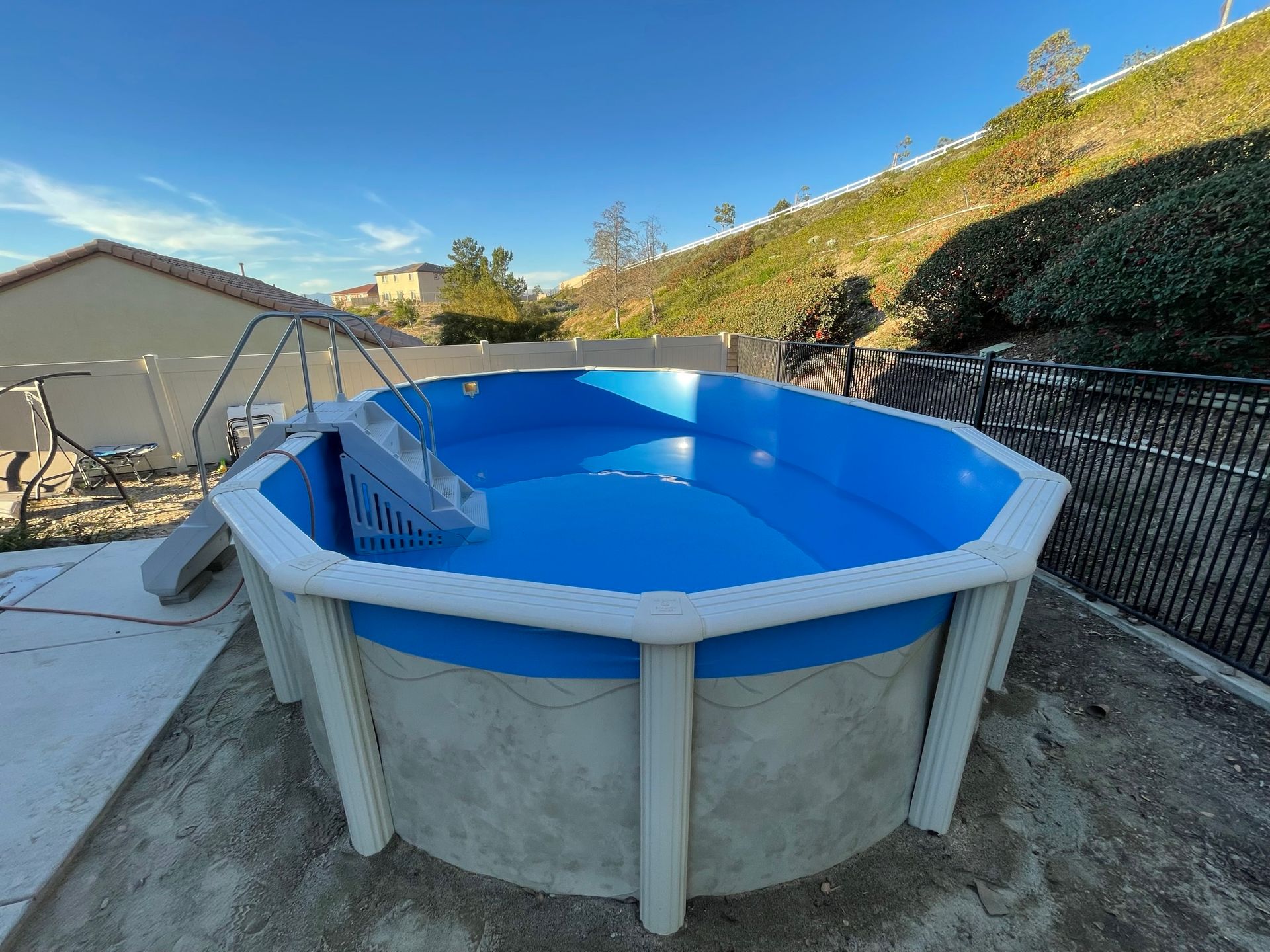 Blue above-ground pool in a backyard, with a ladder and patio area beside a house and hillside.