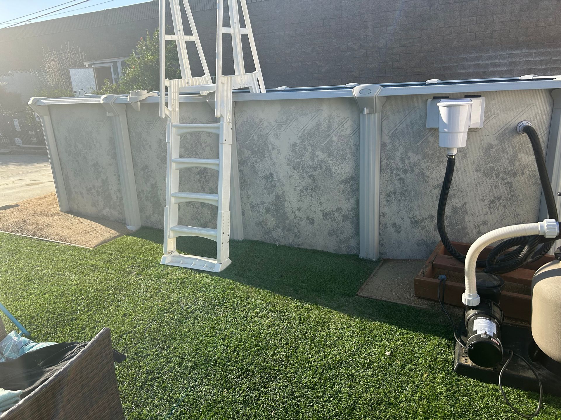 Above-ground pool with white ladder, gray walls, and pool equipment beside a grassy yard.
