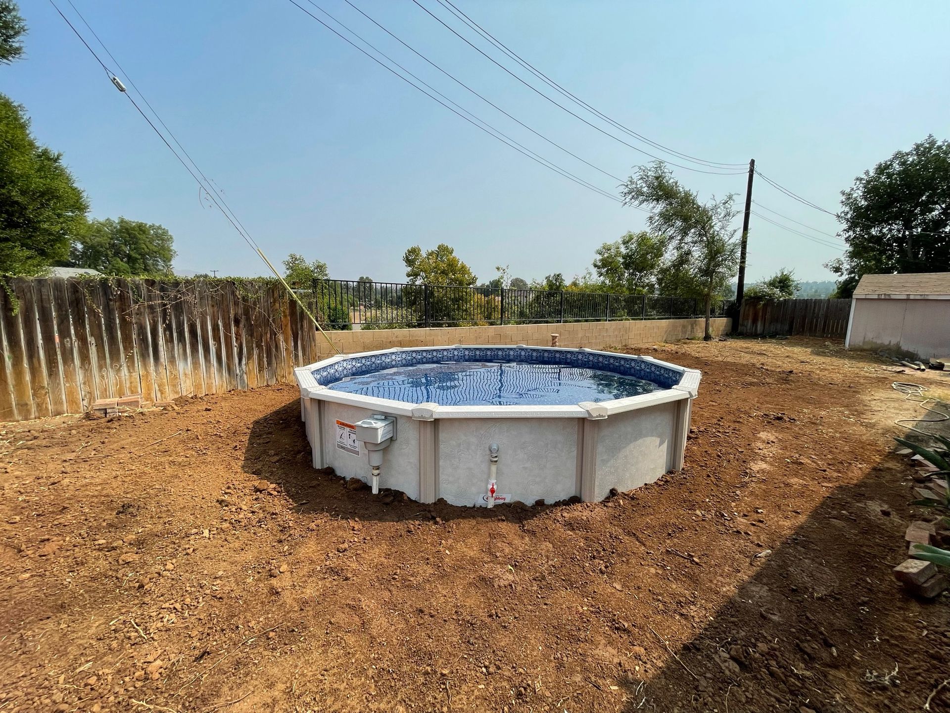 Round above-ground pool in a dry dirt yard with fences and utility poles under a clear sky