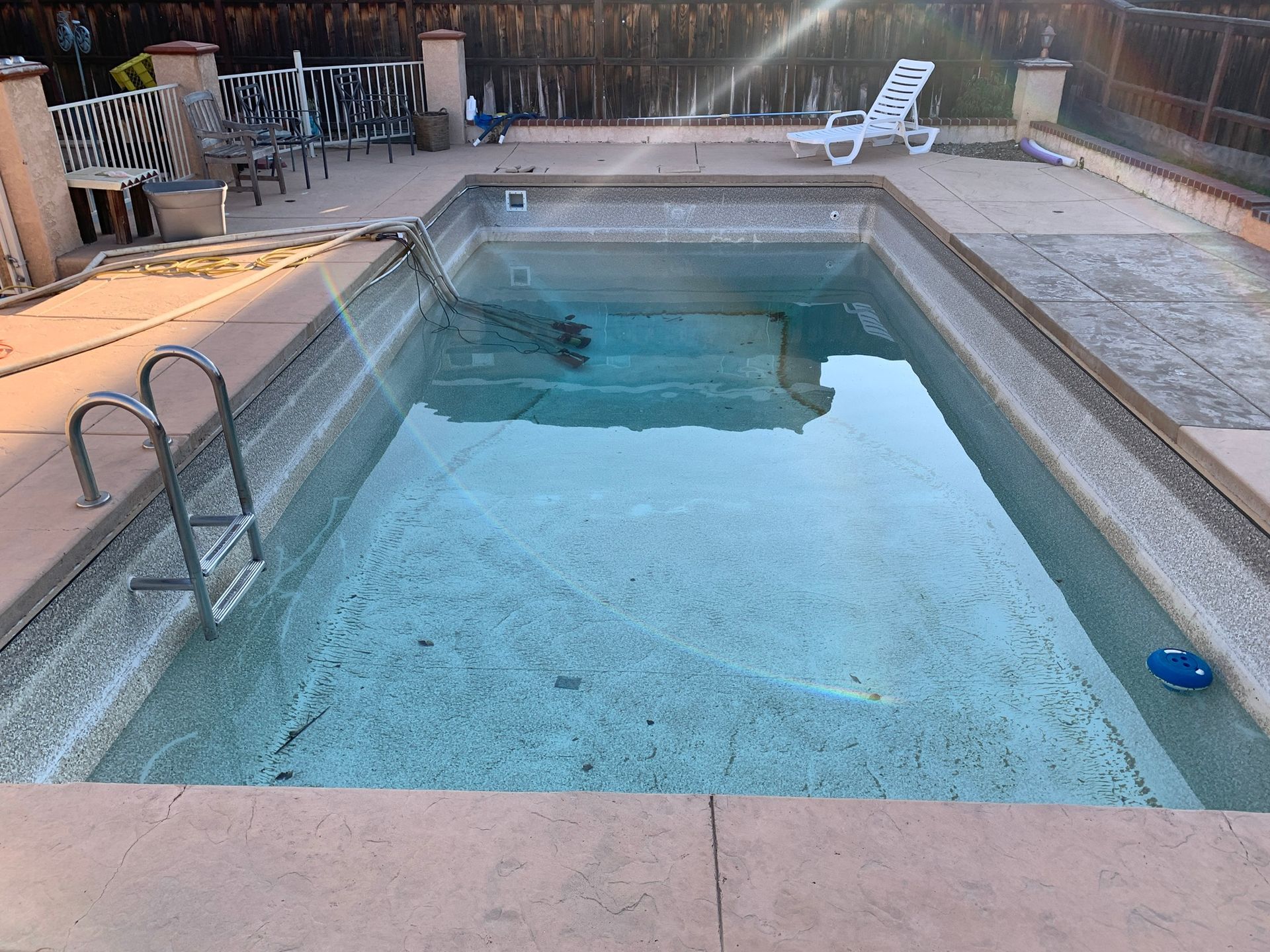 Rectangular backyard swimming pool with ladder, patio chairs, and stone deck at dusk