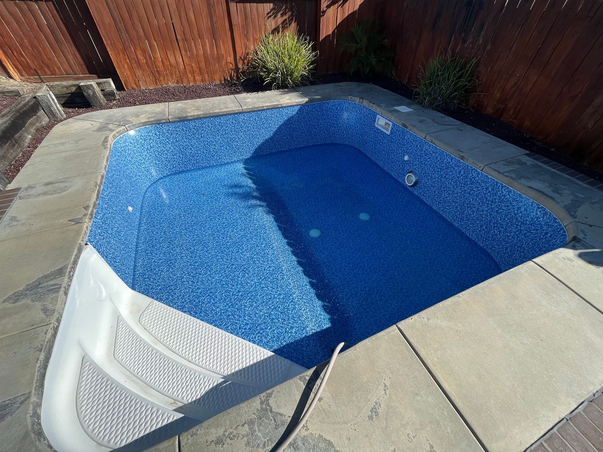 Empty backyard pool with bright blue finish, surrounded by concrete patio and wooden fence.