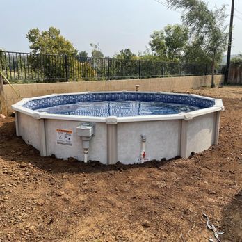 Round above-ground pool with blue water in a dirt yard near a fence and trees