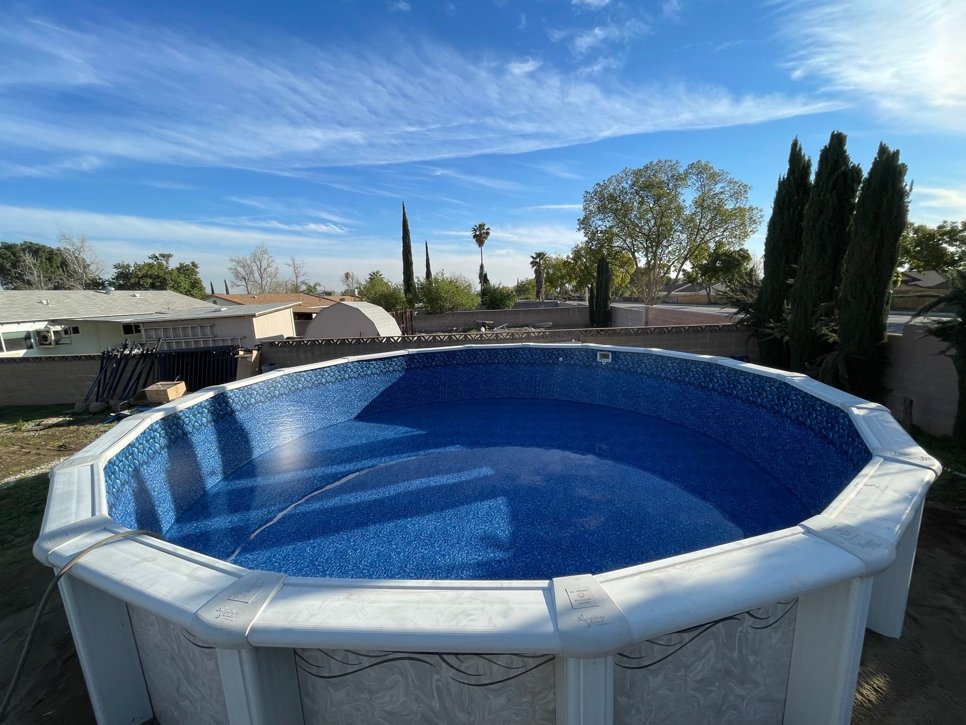 Round above-ground pool with blue liner in a sunny backyard, surrounded by trees and a fence