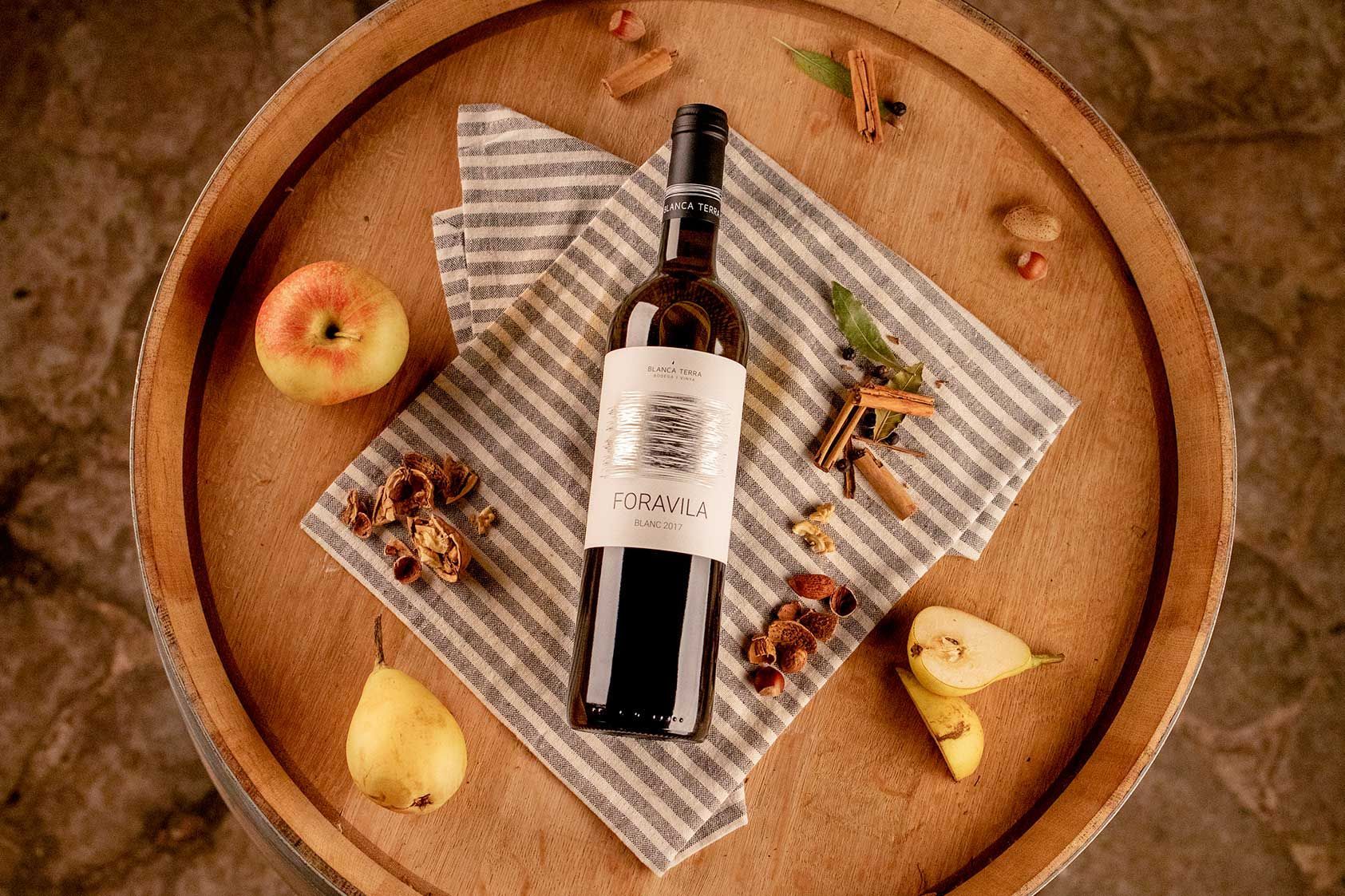 Una botella de vino reposa sobre un barril de madera rodeada de manzanas y peras.