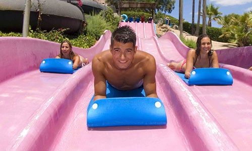 Un hombre y dos mujeres se deslizan por un tobogán de agua rosa en un parque acuático.