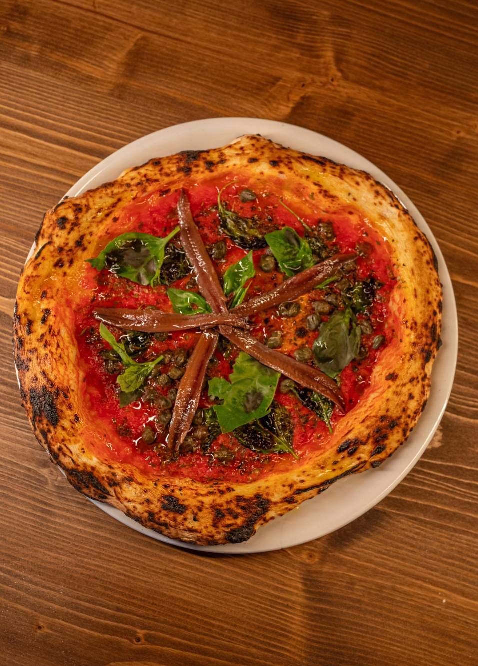 Una pizza con anchoas y espinacas en un plato blanco sobre una mesa de madera.