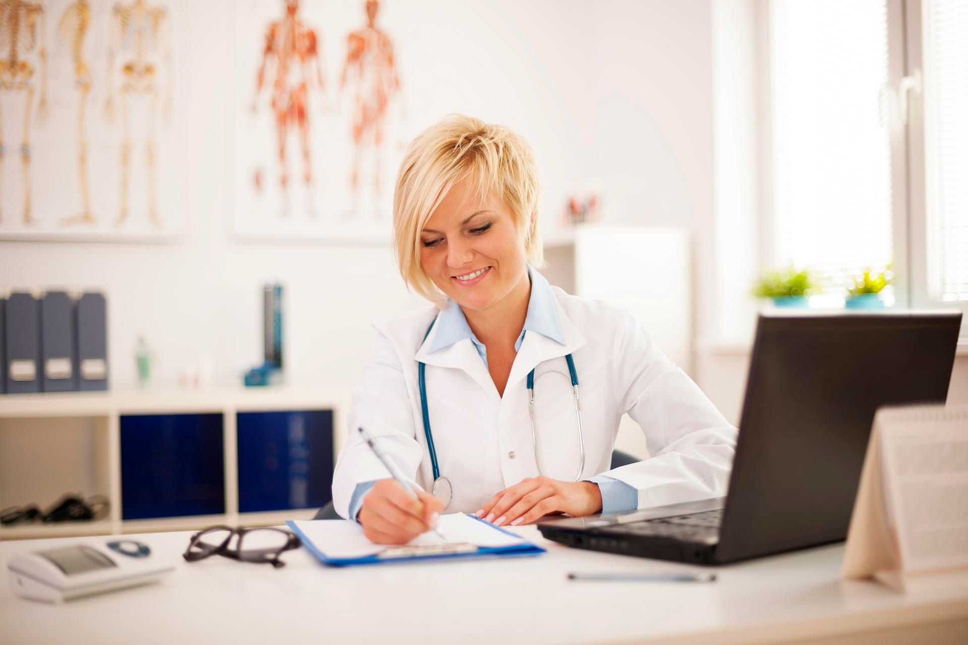 Una doctora está sentada en un escritorio con una computadora portátil y escribiendo en un portapapeles.