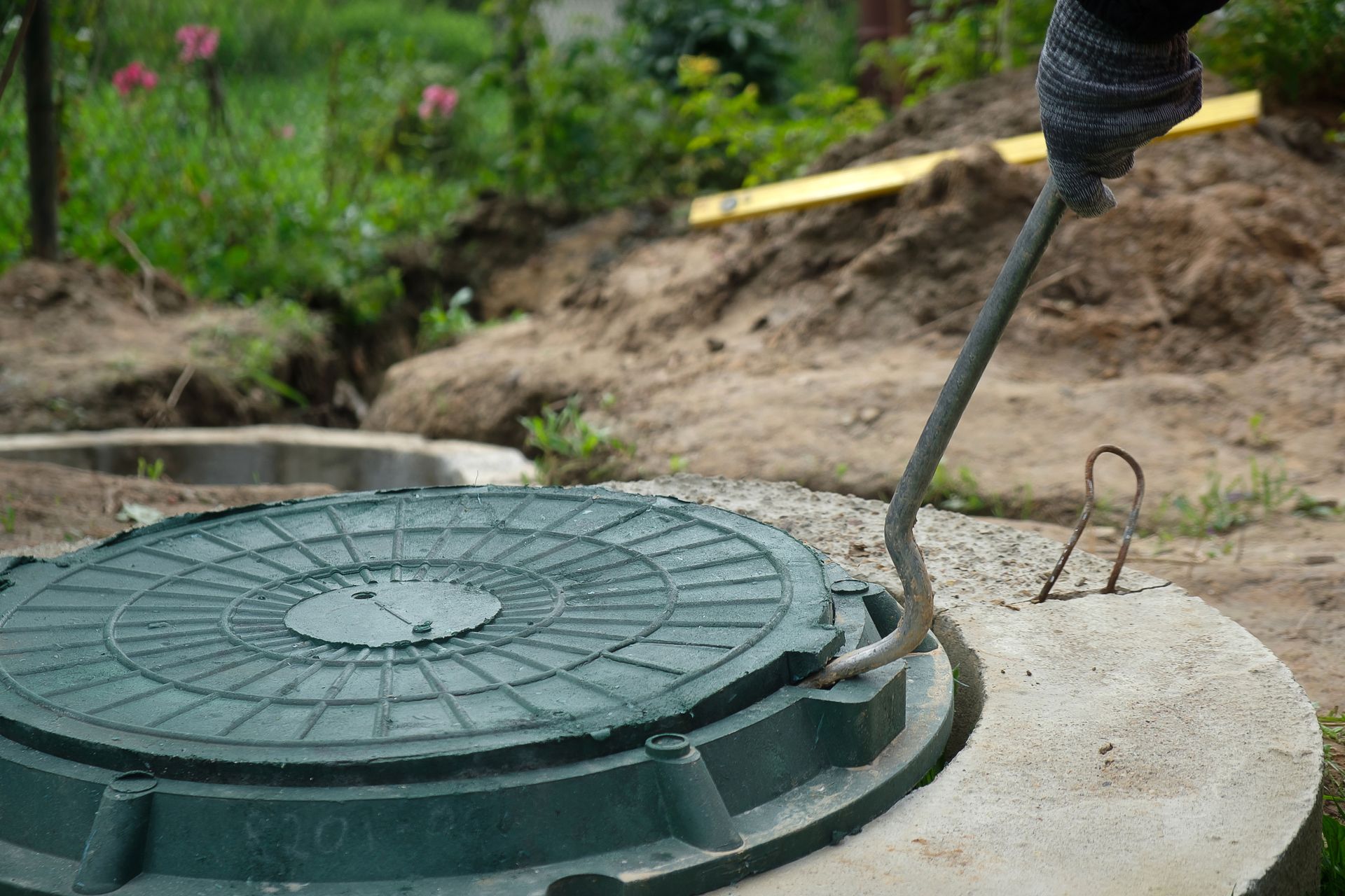 a green manhole cover being lifted with a metal hook by a gloved hand, set in a concrete frame.