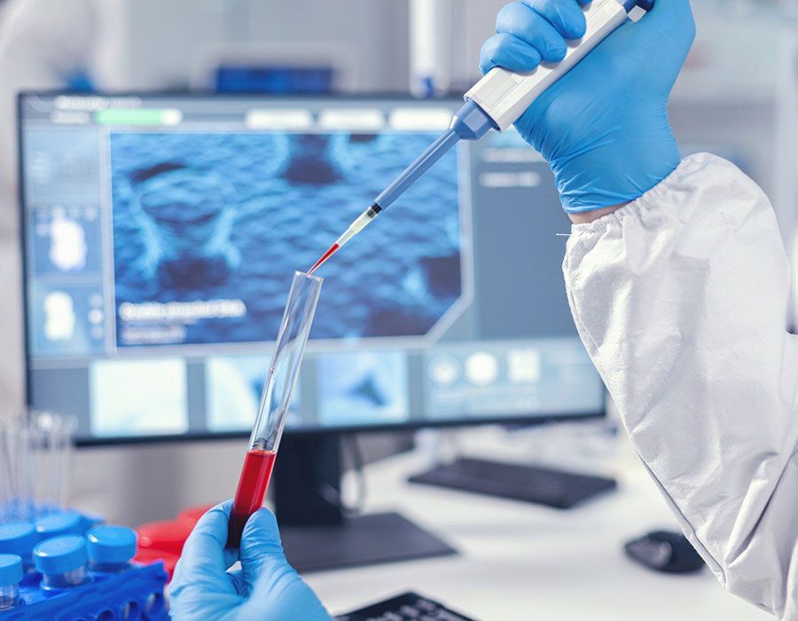 Gloved scientist transferring red liquid between test tubes in a lab with a microscope on a computer screen