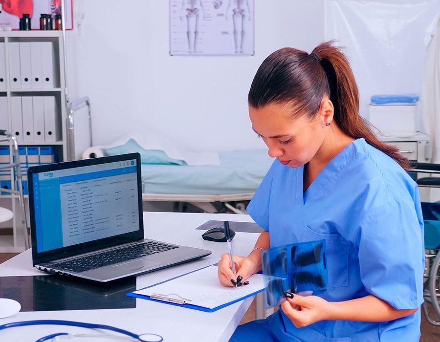 Healthcare worker in blue scrubs writing on a clipboard beside a laptop in a clinic room