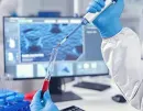 Gloved hands pipetting red liquid in a lab, with a medical scan displayed on a computer monitor.