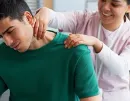A therapist massages a man's neck and shoulders while he leans forward in a treatment room.