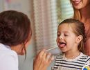 Child opening mouth for a throat check while a doctor uses a tongue depressor; adult stands nearby