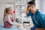 Child and smiling clinician sitting face to face in a bright therapy room, wearing a red clown nose