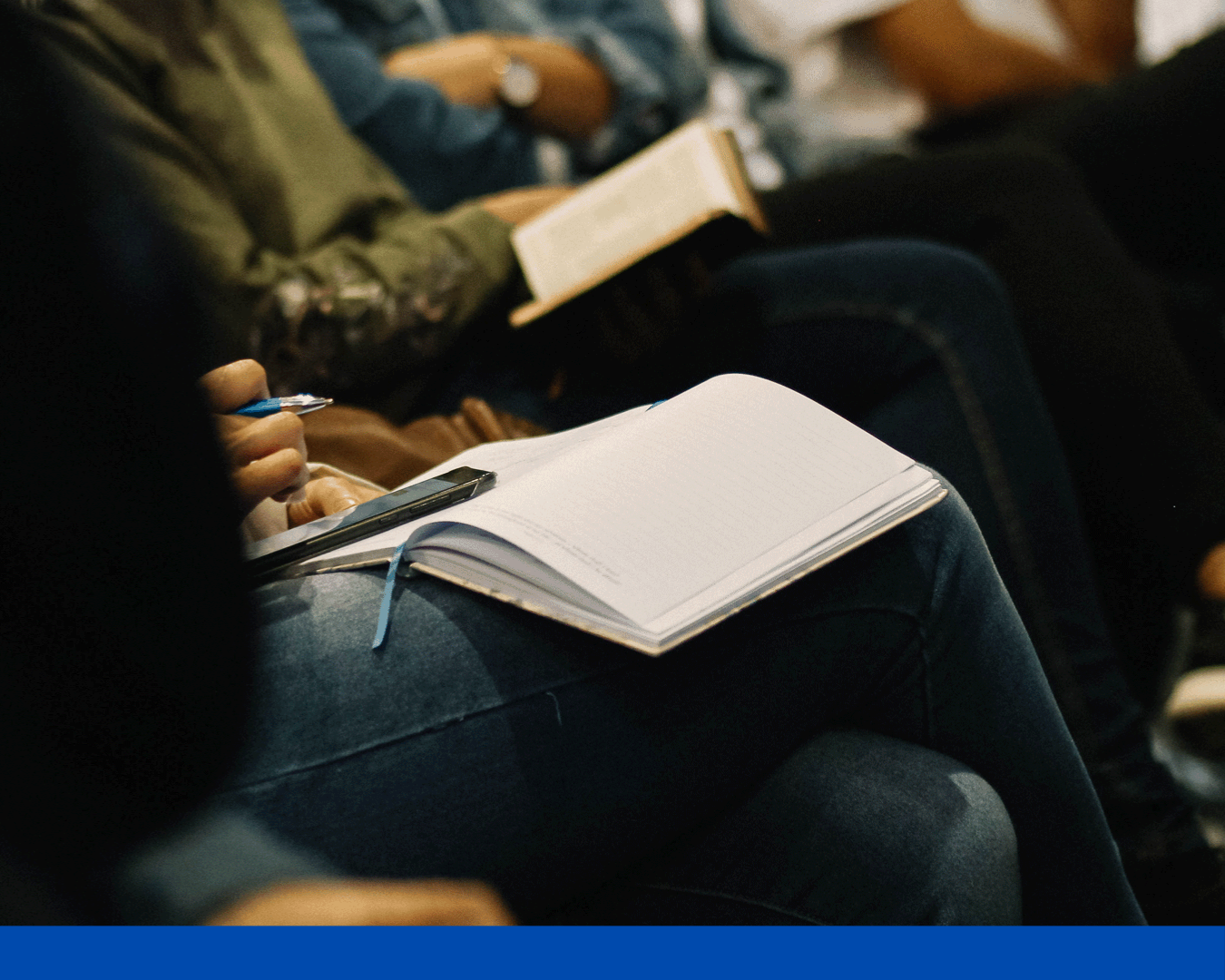 Open notebook on a lap in an audience, with a hand holding a pen and a book nearby