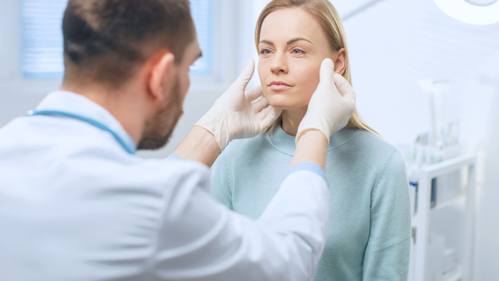 Doctor examines patient’s face in a bright clinic, wearing gloves; patient looks tense.