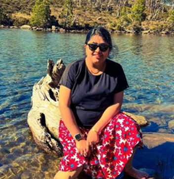 Payel wearing sunglasses and a patterned red skirt sits on a log in shallow, clear water by a mountain shoreline.