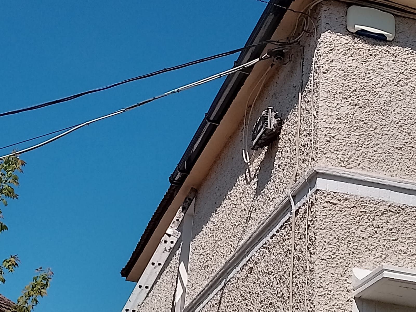 Ladder against a stucco house with wires and a security box on a sunny day.