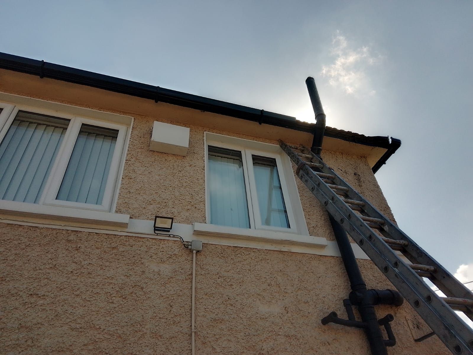 Ladder leaning against a house exterior with two windows. Chimney visible. Sunny day.