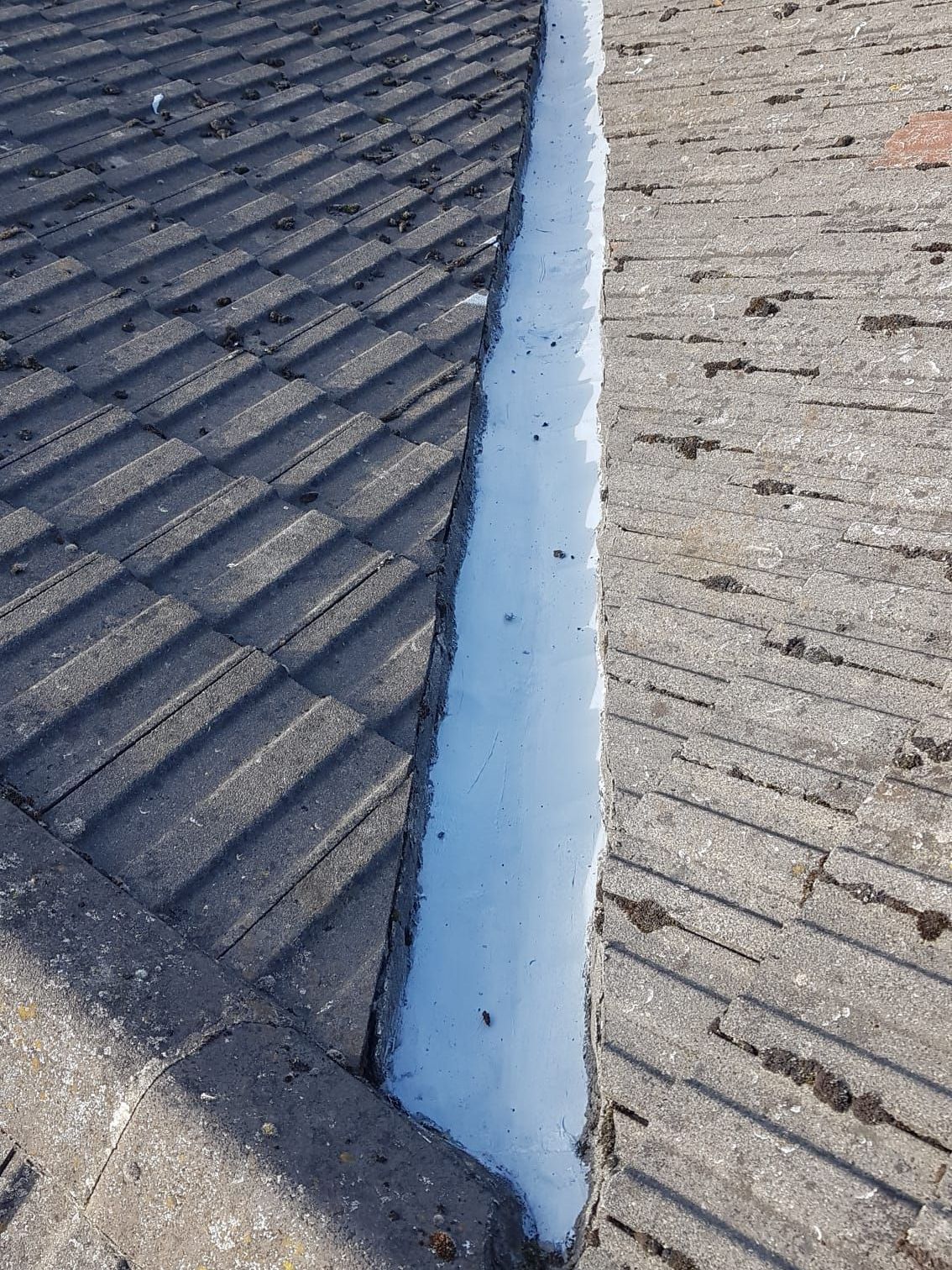 A roof valley with dark gray tiles on one side and light gray roofing on the other; filled with white sealant.