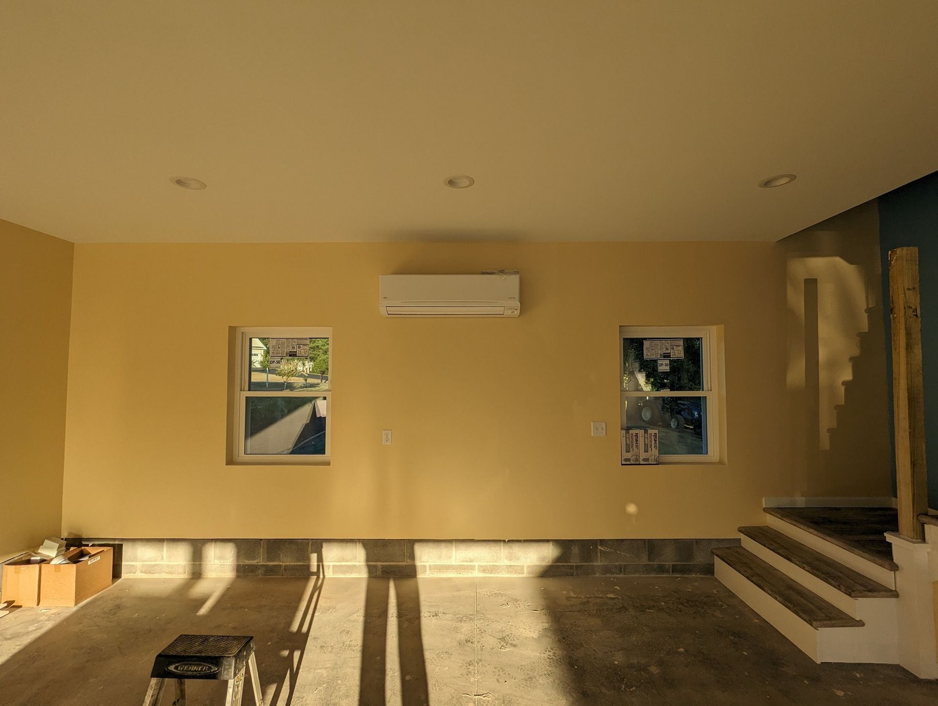 Empty yellow room with two windows, wall AC unit, and stairs on the right, lit by sunlight.
