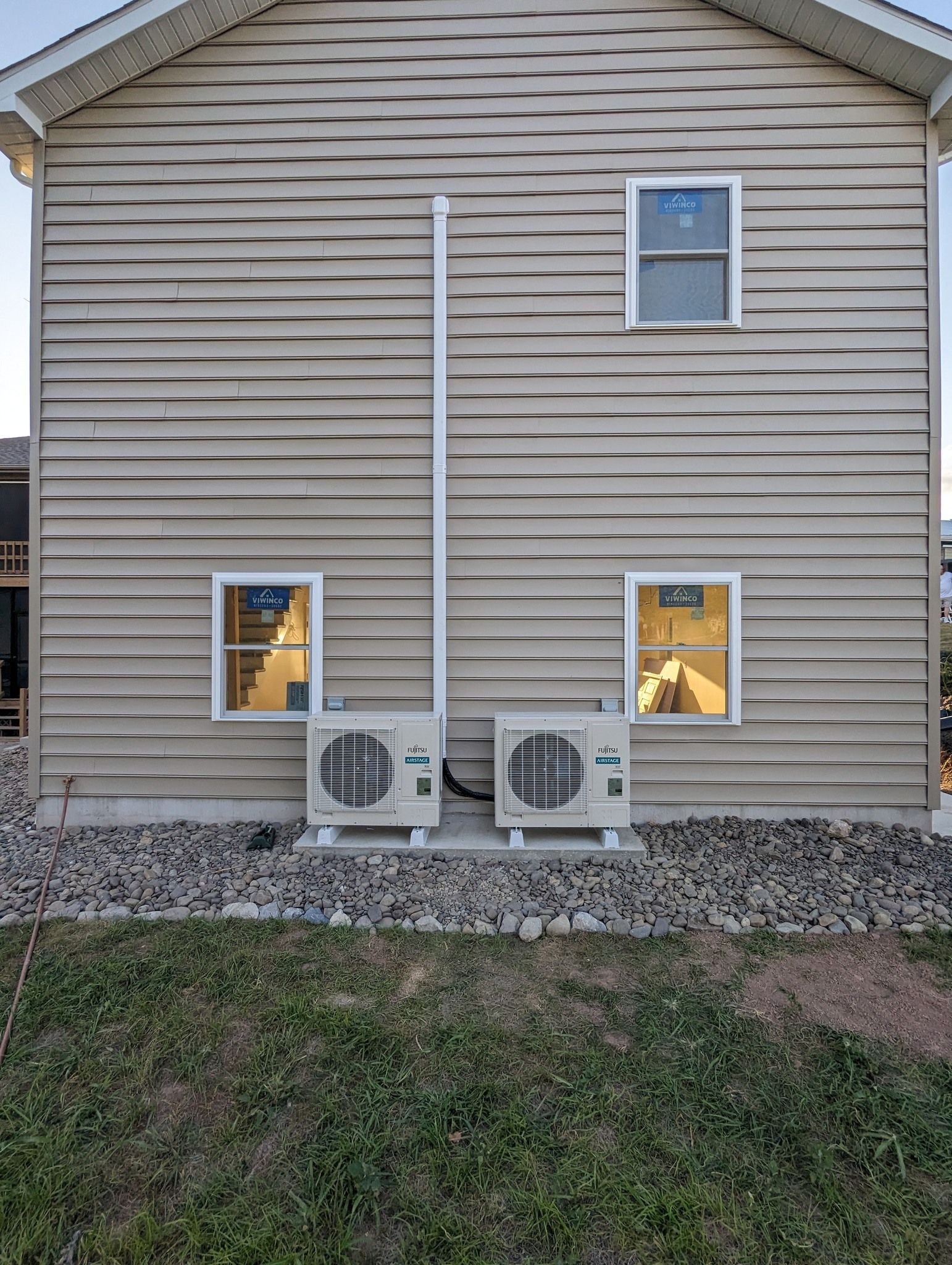 Rear view of a beige house with two AC units, windows, and a gravel strip along the foundation.