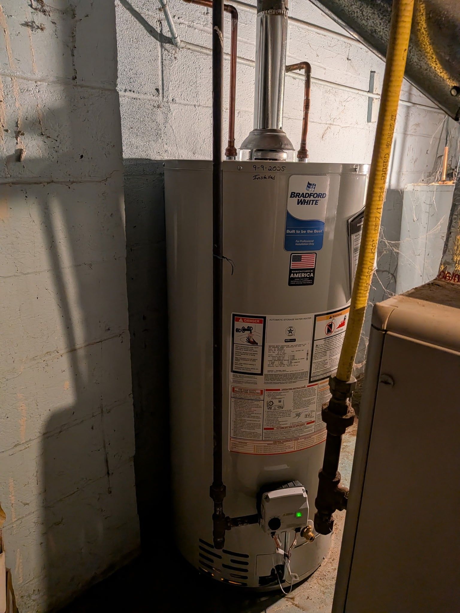 White water heater in a basement beside cinderblock wall, with pipes and a yellow hose attached.