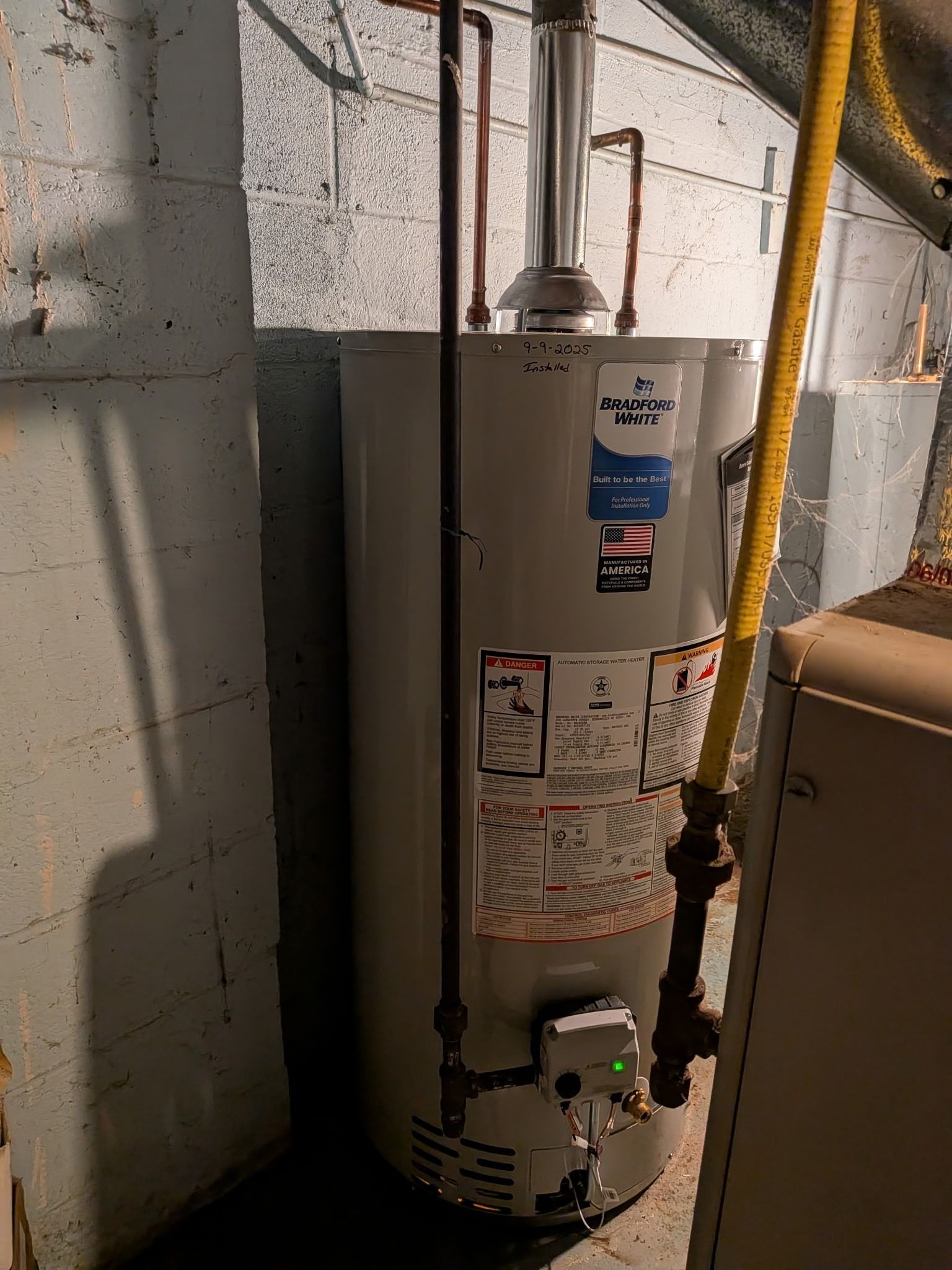 White water heater in a basement utility area with pipes and labels on a cinder block wall