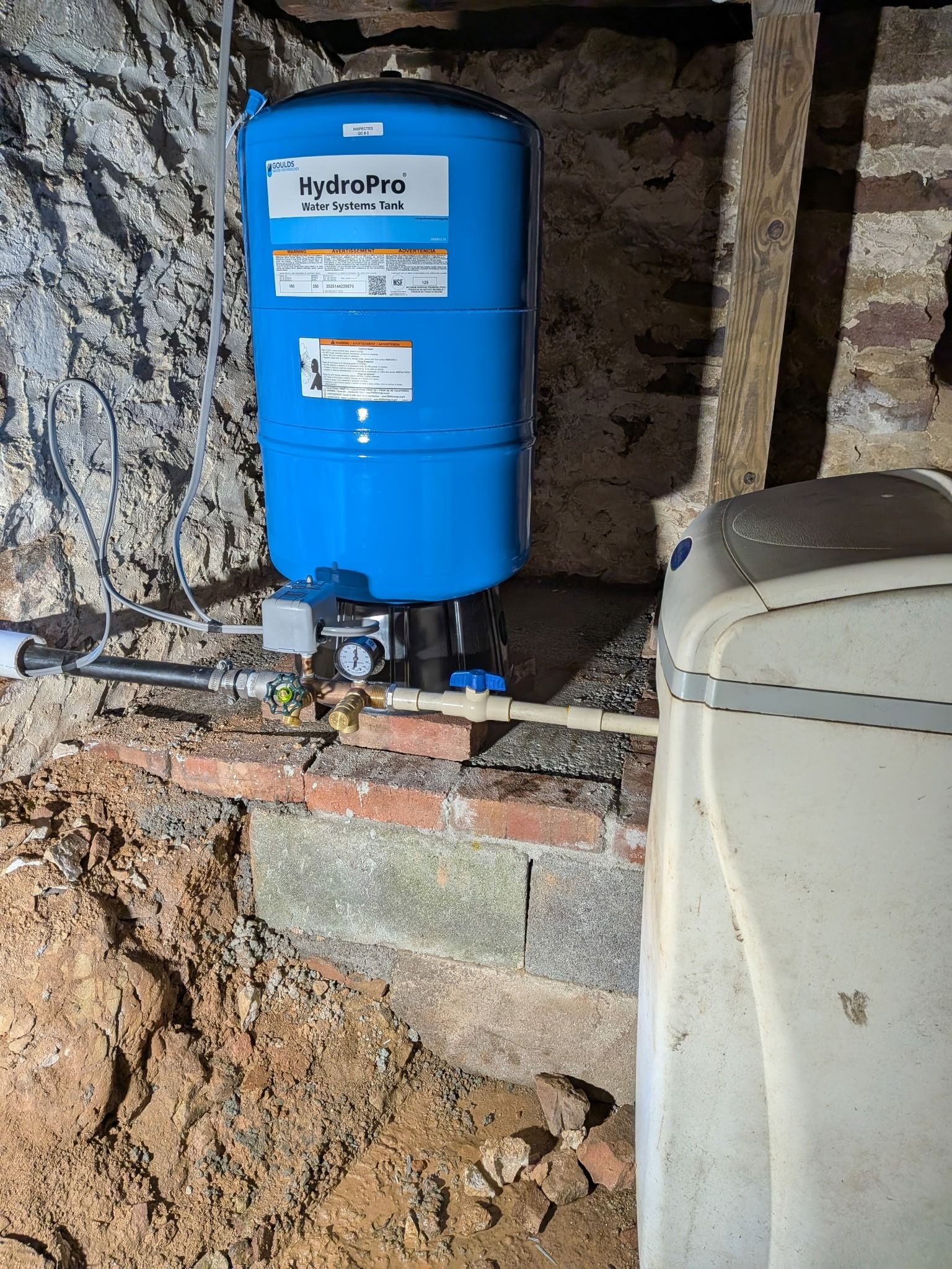 Blue water heater tank installed in a rough stone utility space with exposed pipes and brick support
