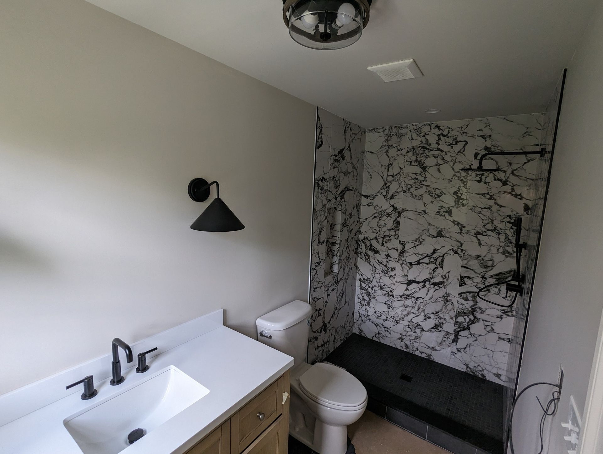 Small bathroom with white sink vanity, toilet, black wall sconce, and marble-patterned shower wall.