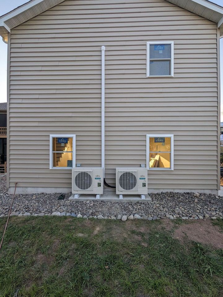 Rear exterior of beige house with two ground-level windows, two AC units, and a vertical pipe or conduit on the wall