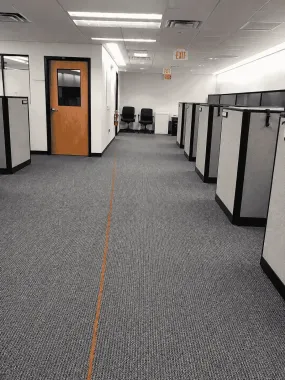 Office hallway with gray carpet, cubicles on both sides, and a wooden door at the end.