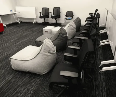 Meeting room with gray bean bags, black chairs, whiteboard, and desks arranged along the walls.
