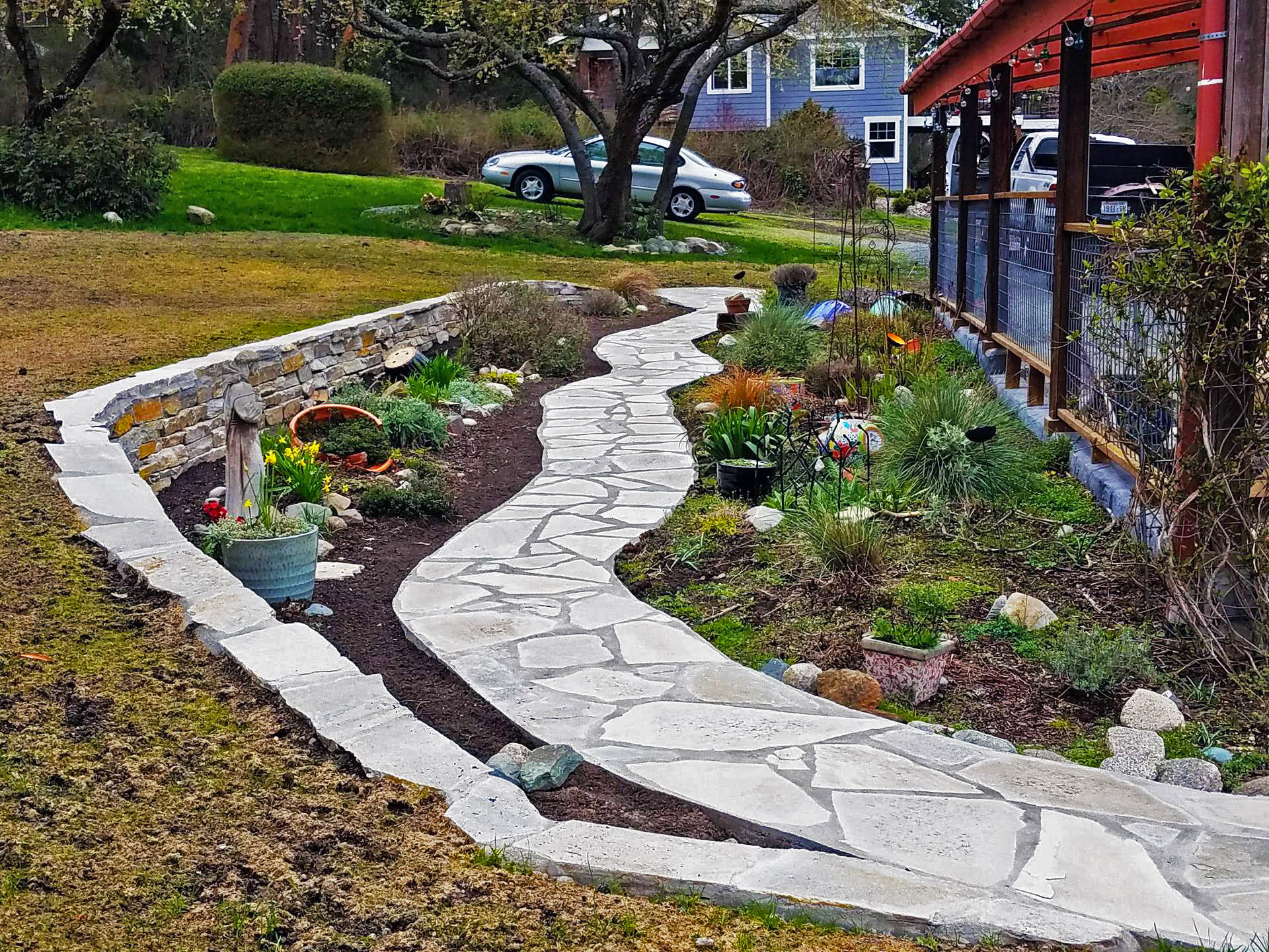 Flagstone walkway winds through a garden with a retaining wall, leading to a house with a red roof.