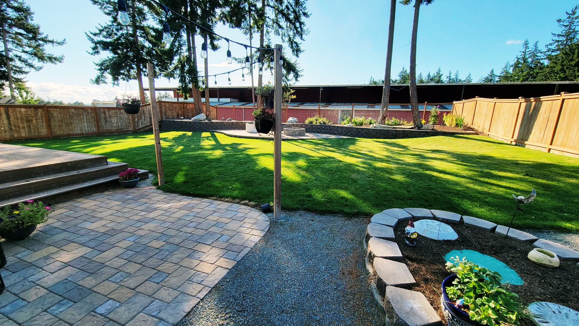 Backyard with green lawn, stone patio, gravel path, and flower beds. String lights hang between trees.