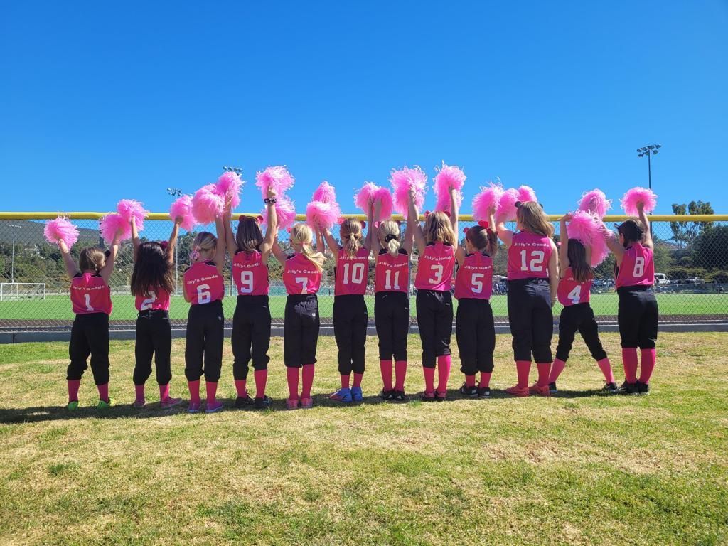 Softball team in pink jerseys holding pink pom-poms on a field under a blue sky