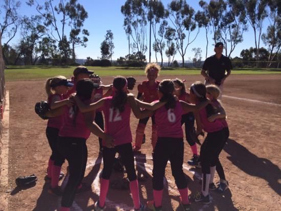 Kids in pink jerseys huddle on a baseball field, with a coach standing nearby.
