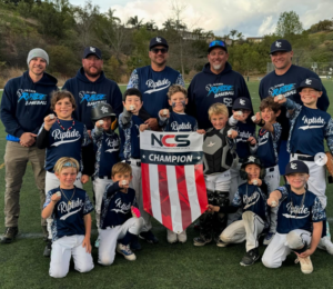 Youth baseball team posing with an NCS Champion banner on the field; coaches stand behind them.