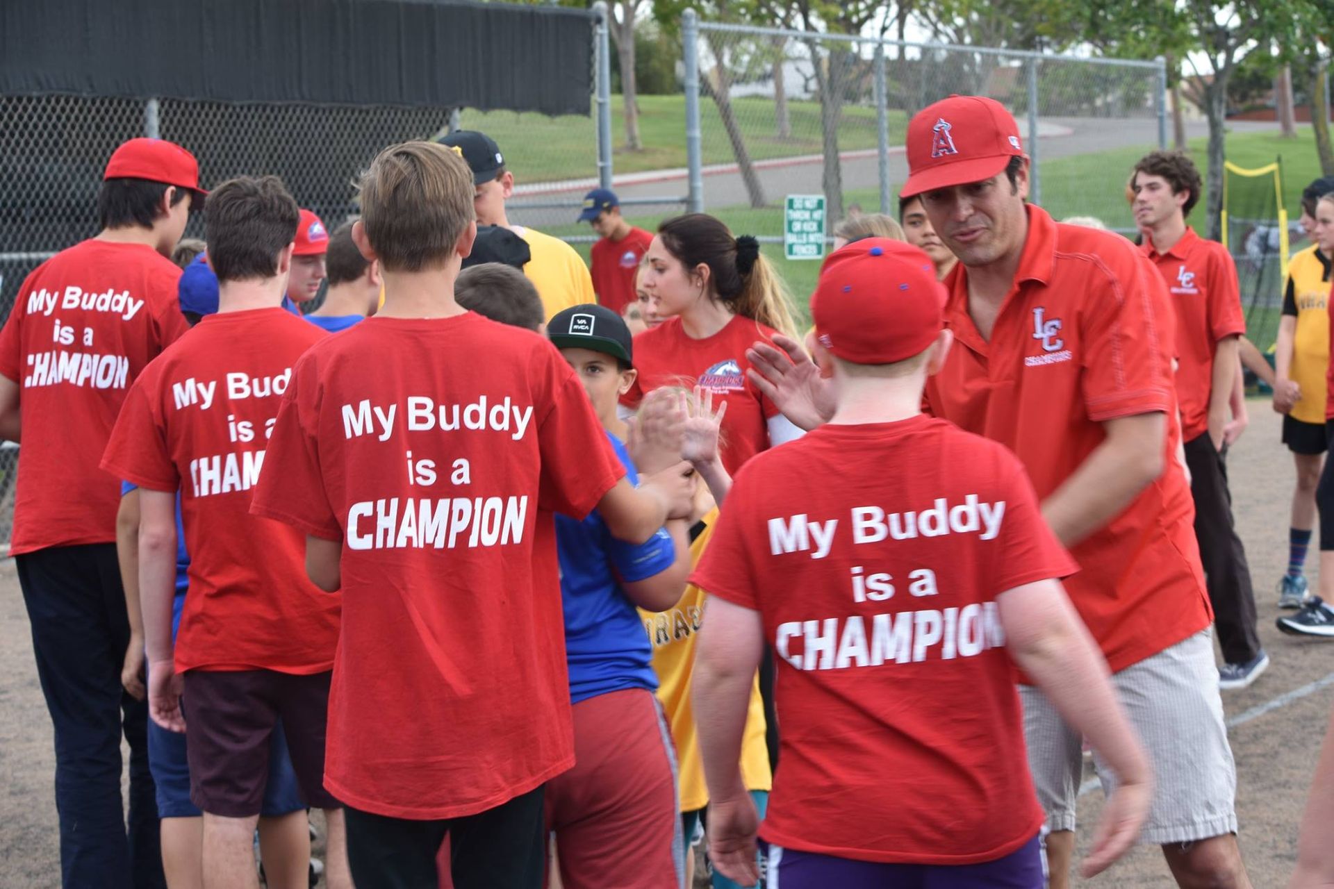 People in red shirts and caps with “My Buddy is a CHAMPION” printed on the back, gathered outdoors.