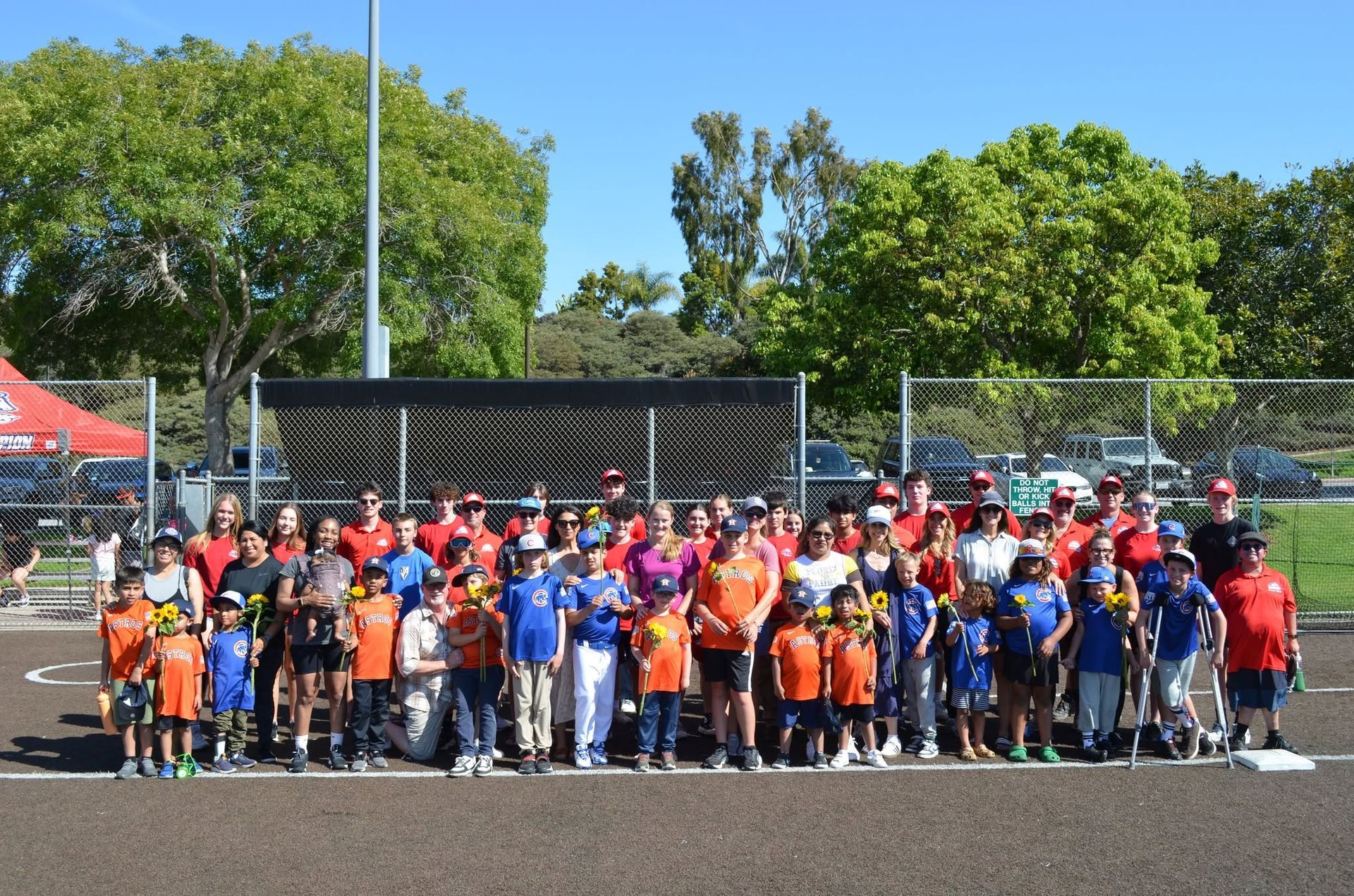 Large group posing on a baseball field, many in orange shirts, with a red tent and trees in the background