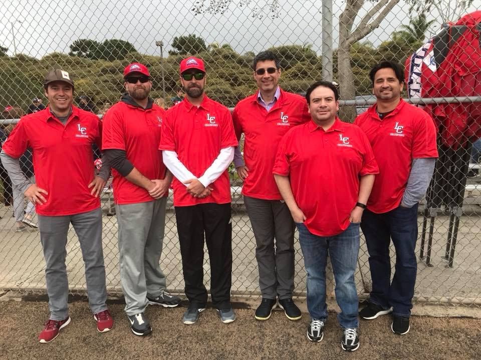Six people in red shirts standing outdoors in front of a chain-link fence