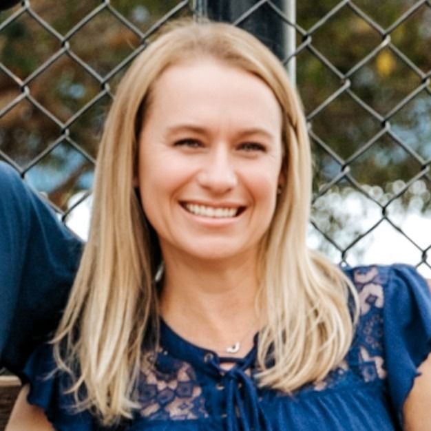 Smiling blonde woman in a blue top outdoors with fence behind her
